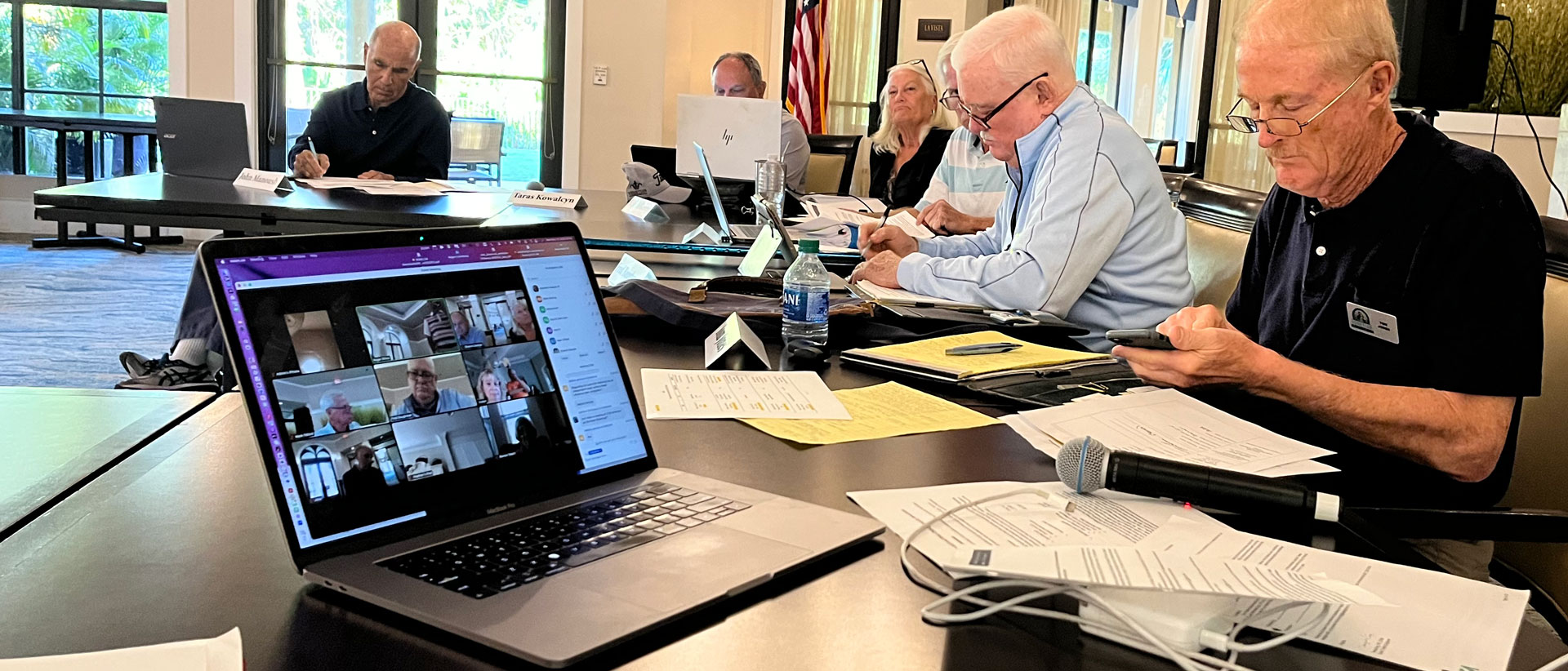 Board meeting with six people sitting around a table and a Macbook with several other participants joining the meeting remotely